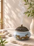 Quemador de incienso de cerámica azul con grabados de dragón imperial y rejilla metálica decorativa, rodeado de elementos zen y velas.